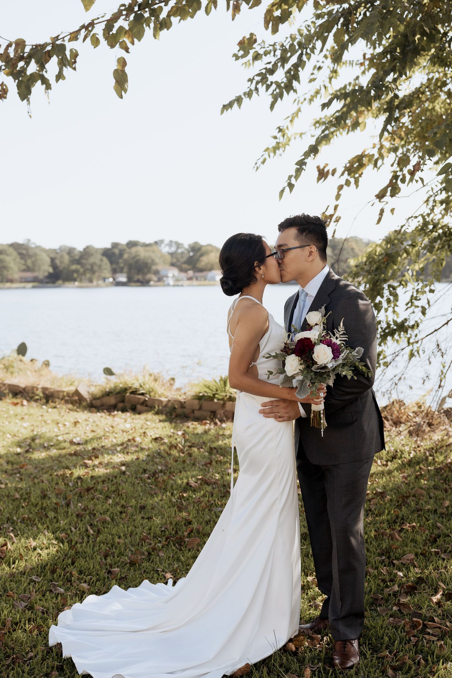 A romantic wedding couple shares a tender moment by a lakeside under overhanging tree branches on a sunny autumn day.