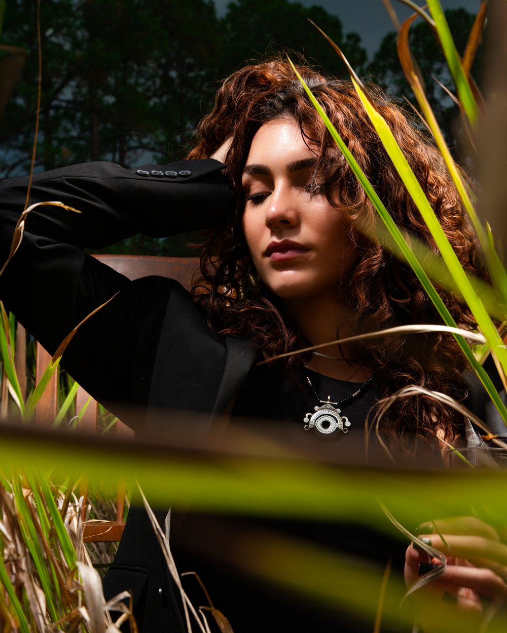 Artistic portrait through tropical foliage showing curly hair and black clothing against a natural outdoor backdrop.