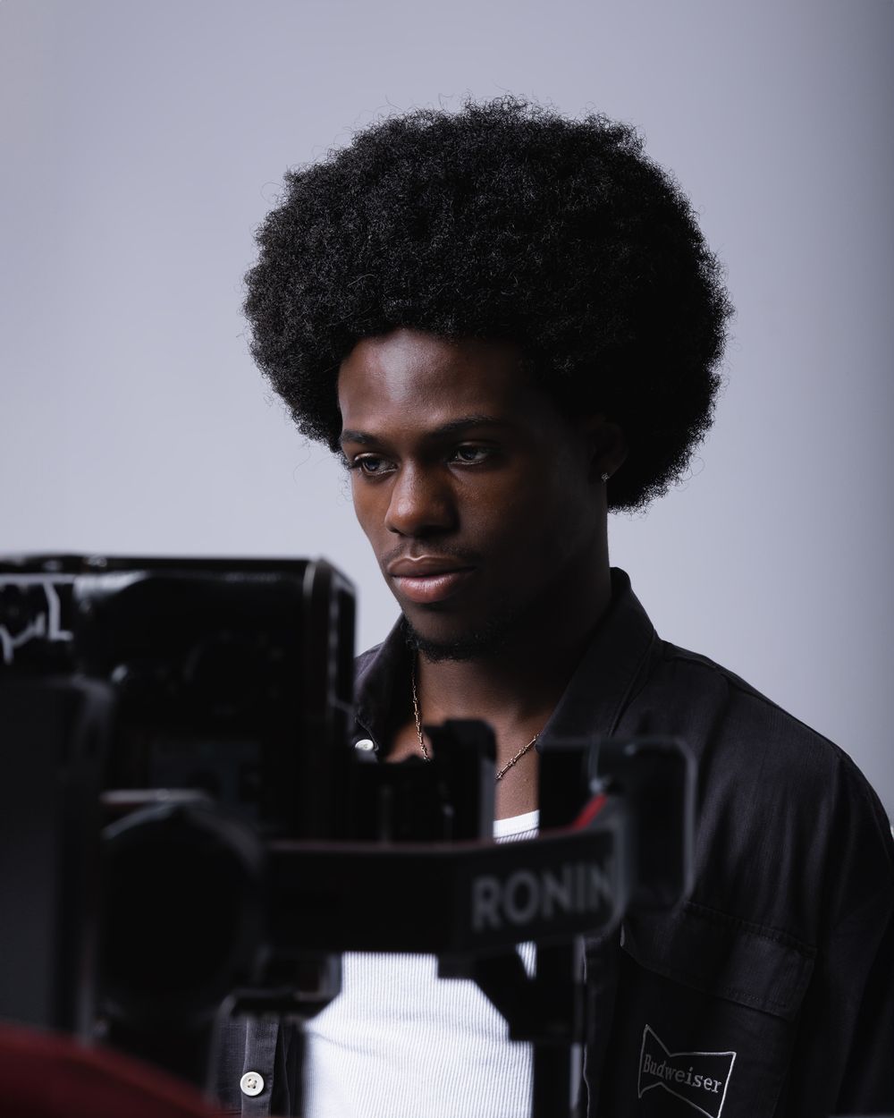 Camera operator wearing black shirt with natural afro hairstyle working with Ronin camera equipment against light background.