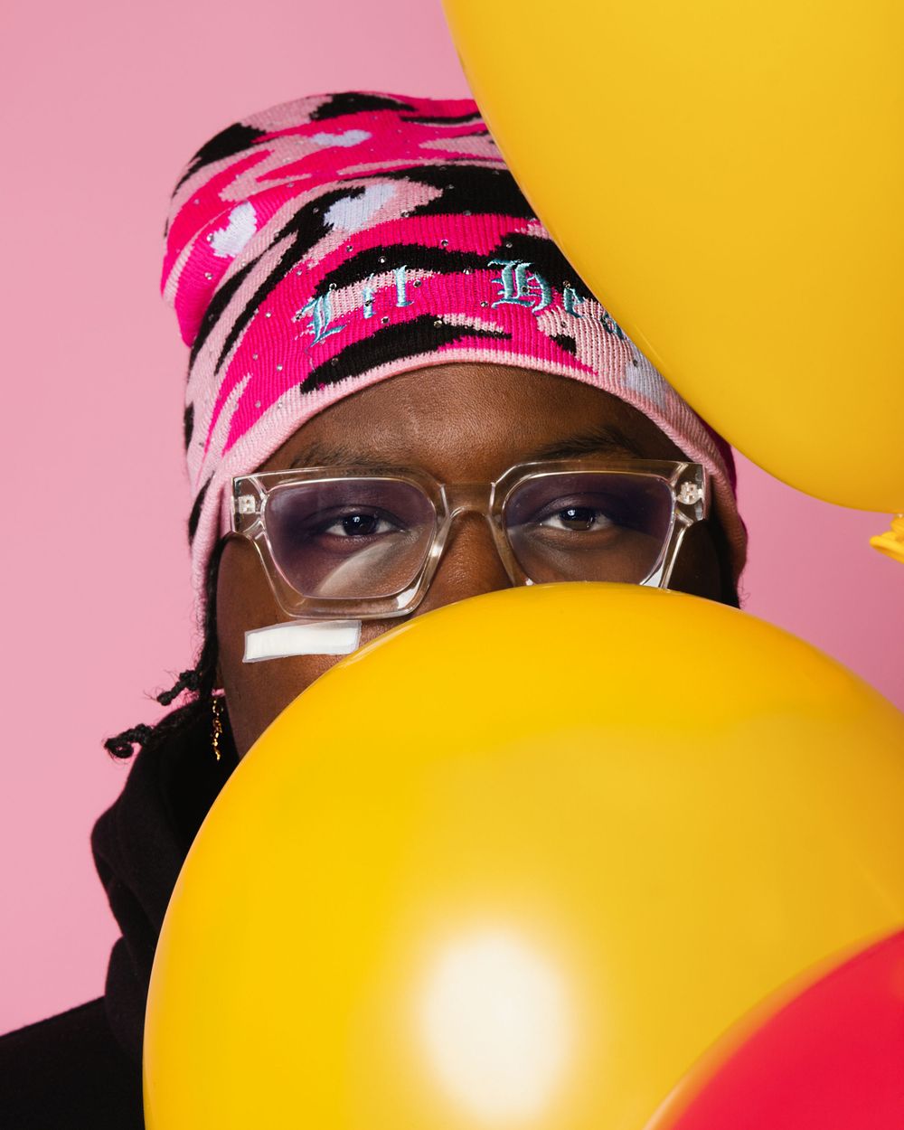 Stylish person wearing clear glasses and pink patterned headwear poses with bright yellow balloons against pink background.