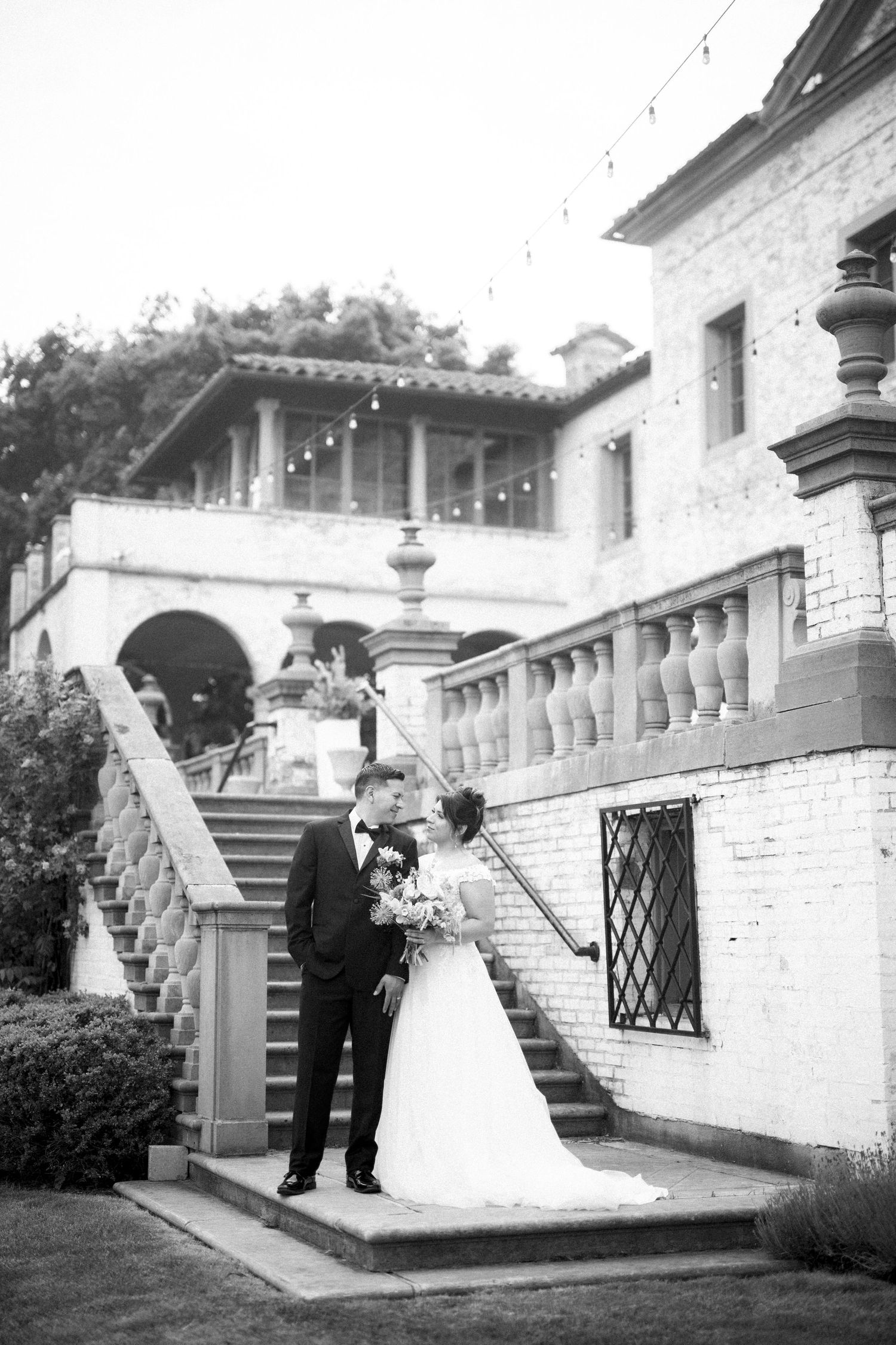 A black and white wedding photo showing a couple standing on elegant stone stairs of a Mediterranean-style villa.