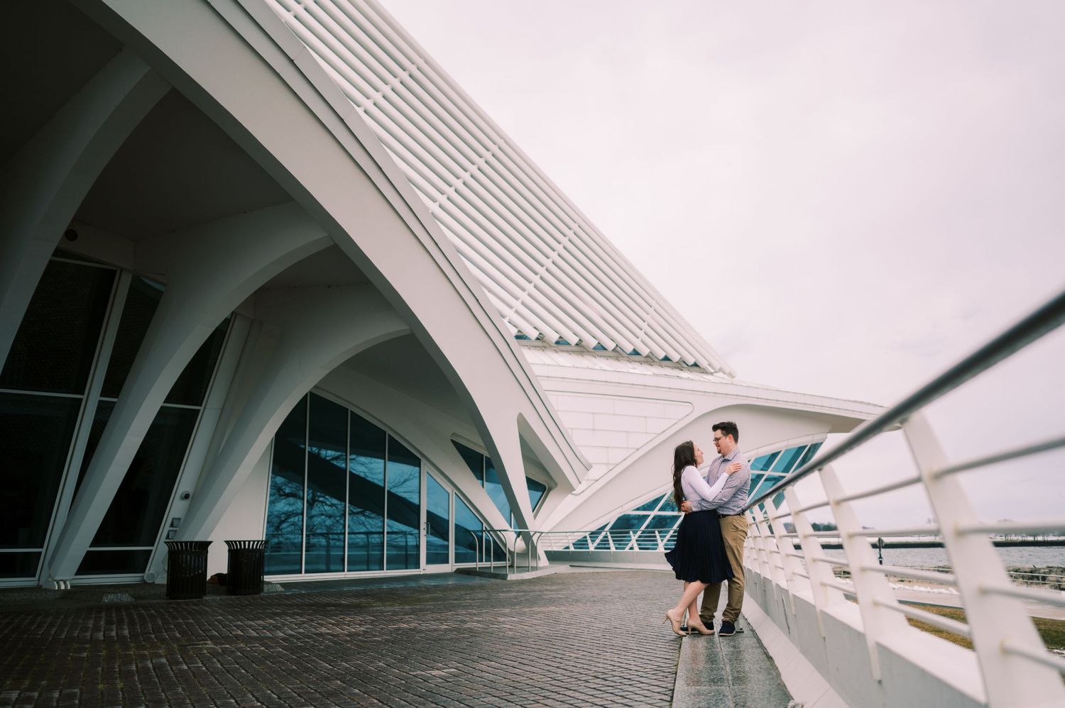 A couple embraces under the sweeping modern architecture of a white arched building with glass windows.