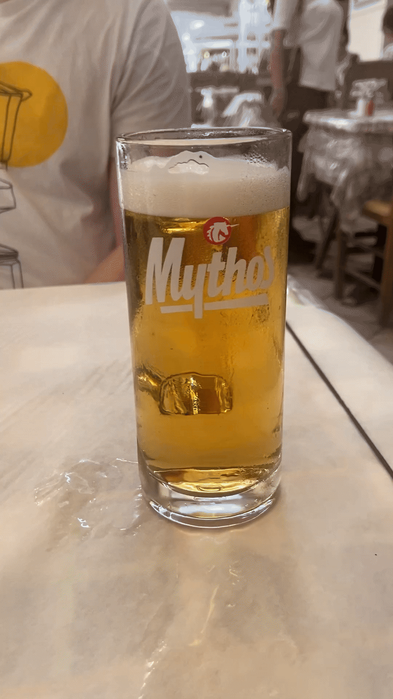 A tall glass of golden Mythos beer with white foamy head sits on a table in what appears to be a restaurant or bar.