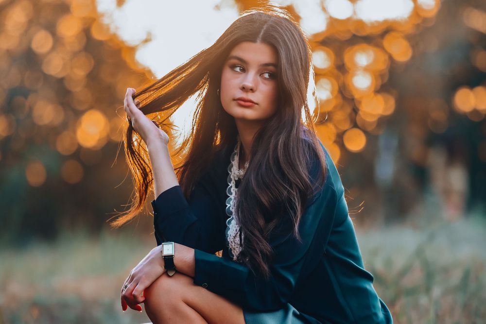 Person in black coat poses outdoors against blurred bokeh lights during golden hour creating moody autumn atmosphere.