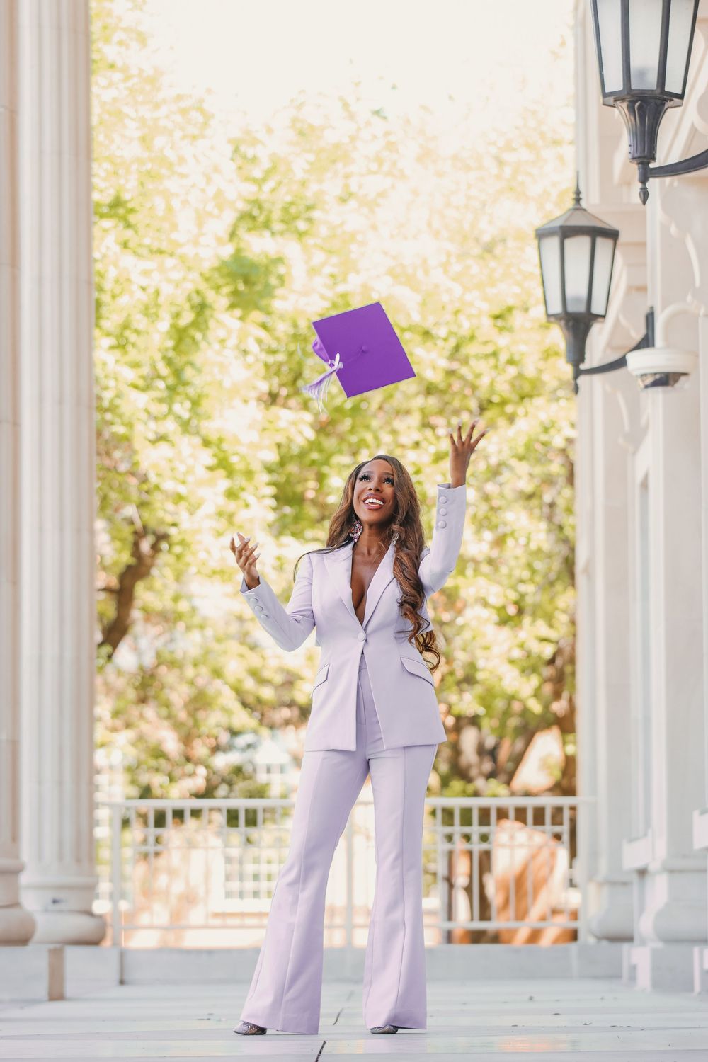 A person in a lavender pantsuit celebrates by tossing a graduation cap in the air near white columns.