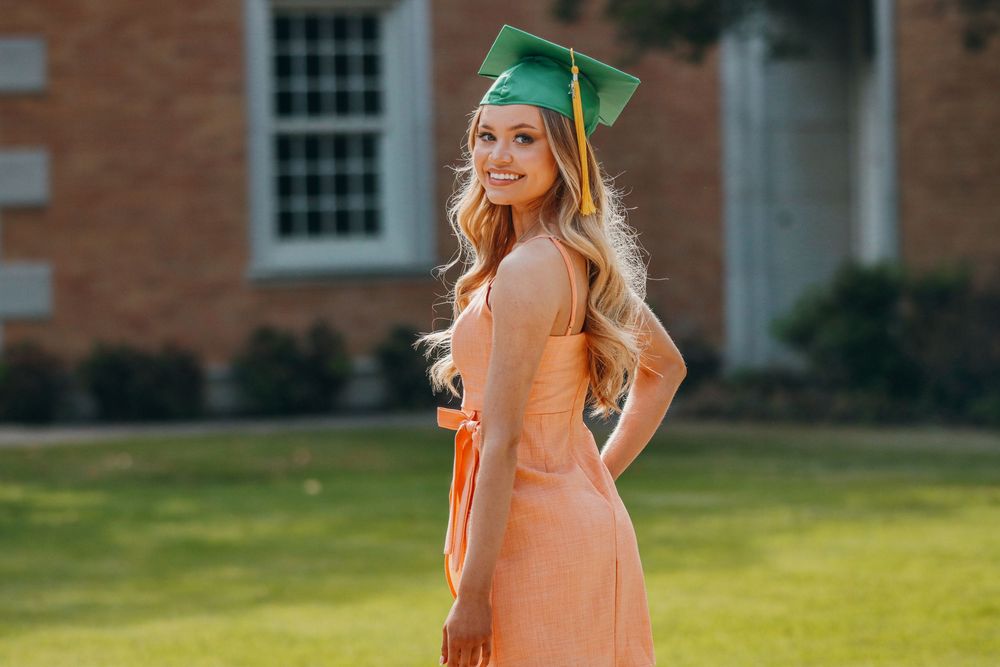 Graduate in a peach-colored dress and green cap poses on a sunny day in front of a brick building.
