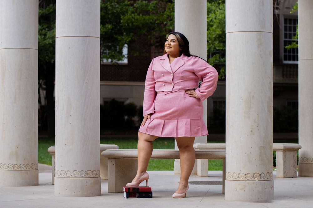 Woman in pink blazer and black shorts poses confidently between white marble columns outdoors.