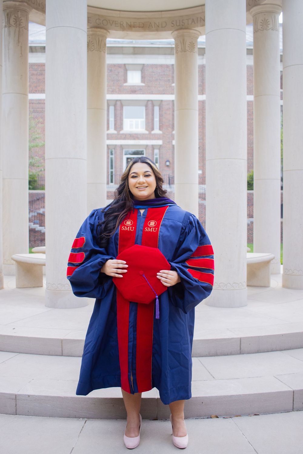 Graduate in navy blue and red academic regalia poses for photos in front of white stone columns at university.