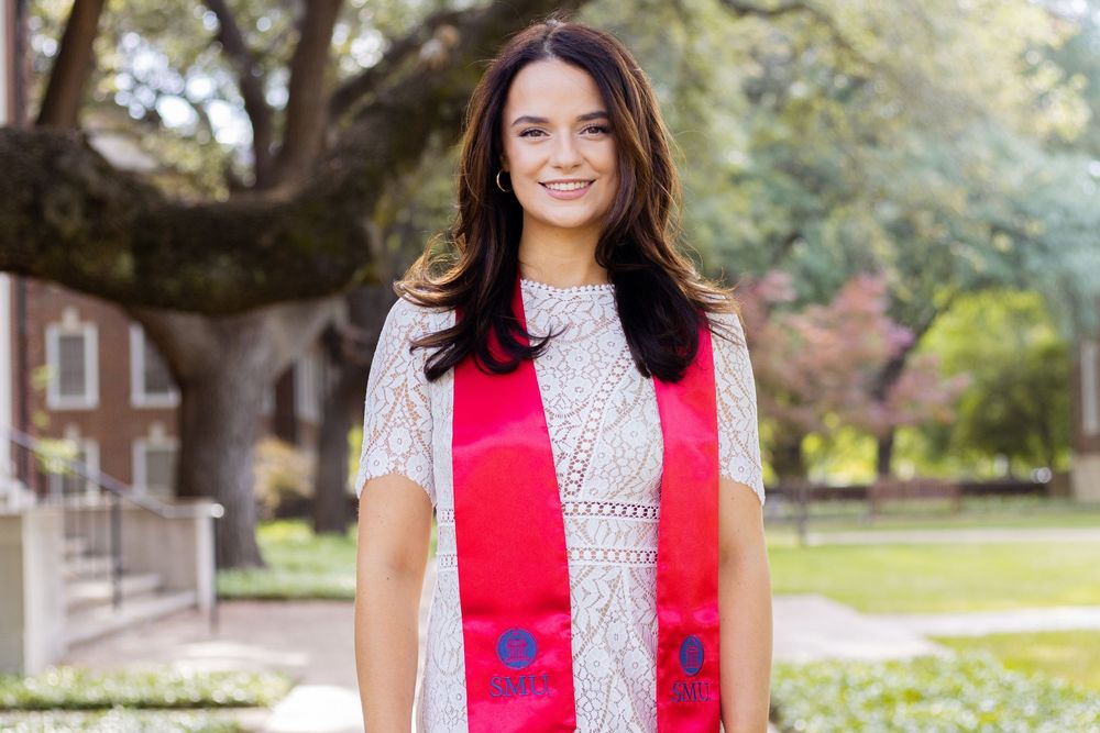 A person in a bright red vest smiles while standing outdoors near brick buildings and trees on a sunny day.