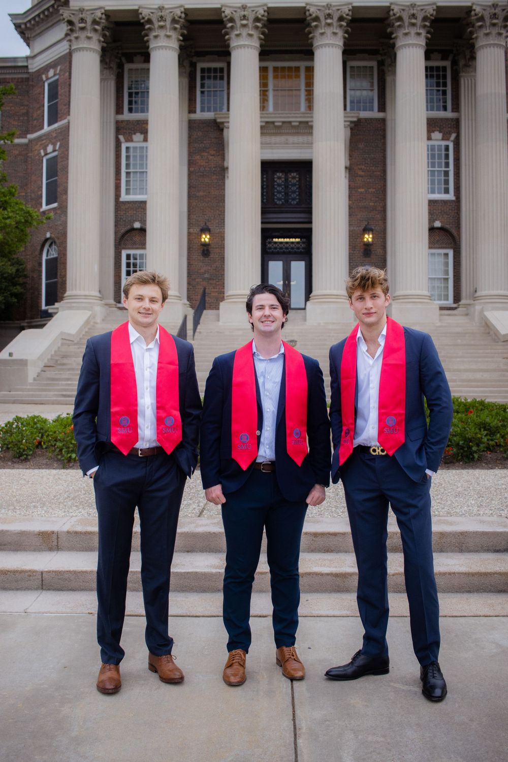 Three graduates in navy suits and red stoles pose together on campus steps in different creative stances.