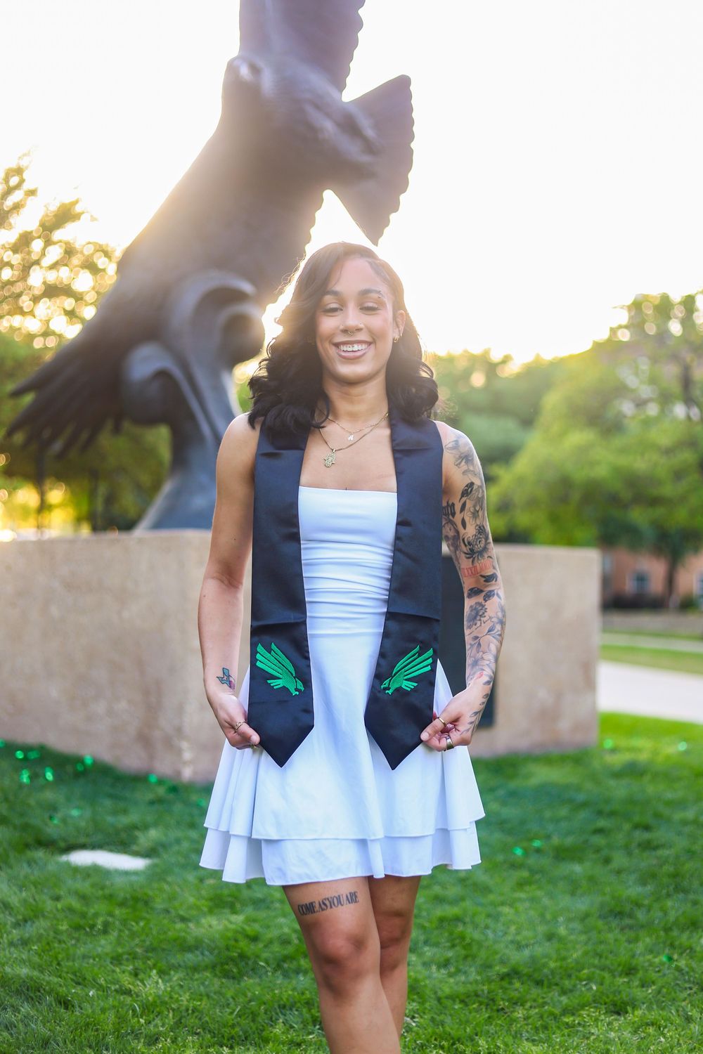 A person wearing a white dress and navy vest with green accents smiles outdoors during golden hour sunset.