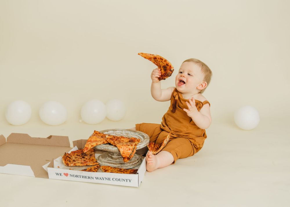 Baby in brown romper eating pizza while sitting on floor with pizza box.