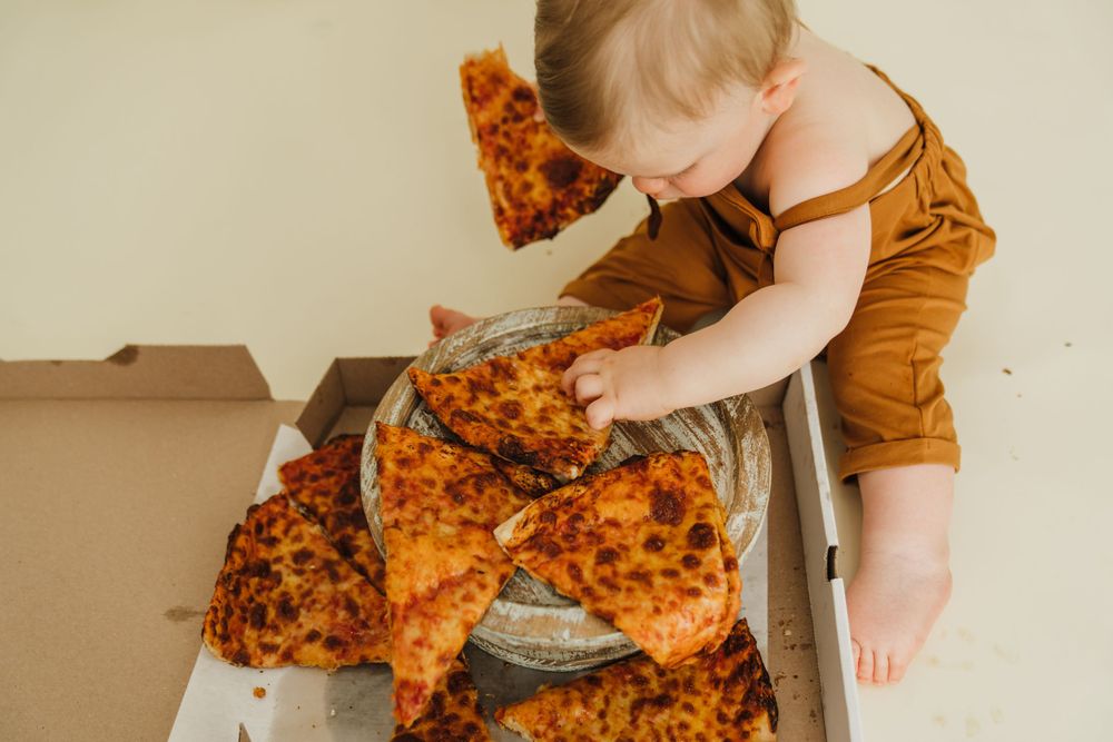 Baby in brow outfit reaching for pizza in cardboard box on light background.