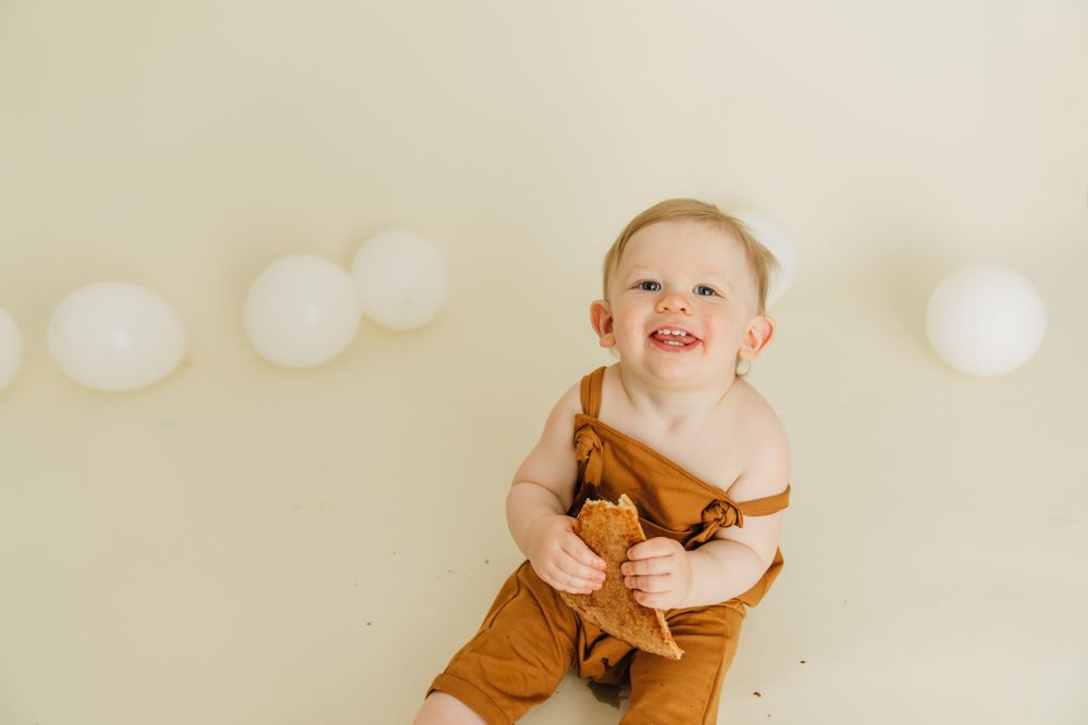 Happy baby in brown romper against cream wall with white balloon decorations.