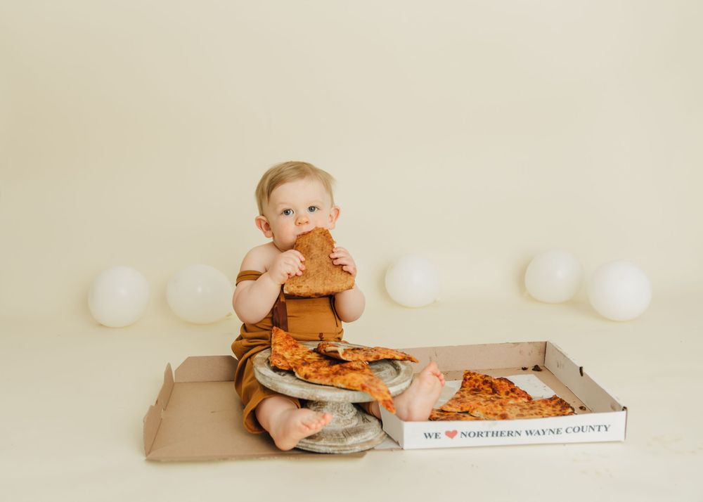 Small child in brown outfit enjoying pizza from delivery box on beige background.