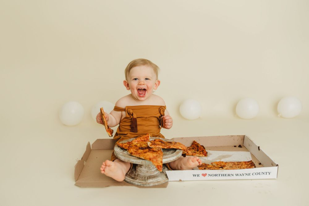 Joyful baby in brow  romper next to pizza box with white decorative balloons.