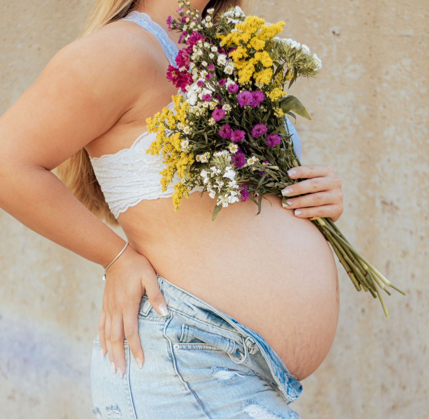 A pregnant belly in a white crop top holding a vibrant wildflower bouquet with yellow and purple blooms.