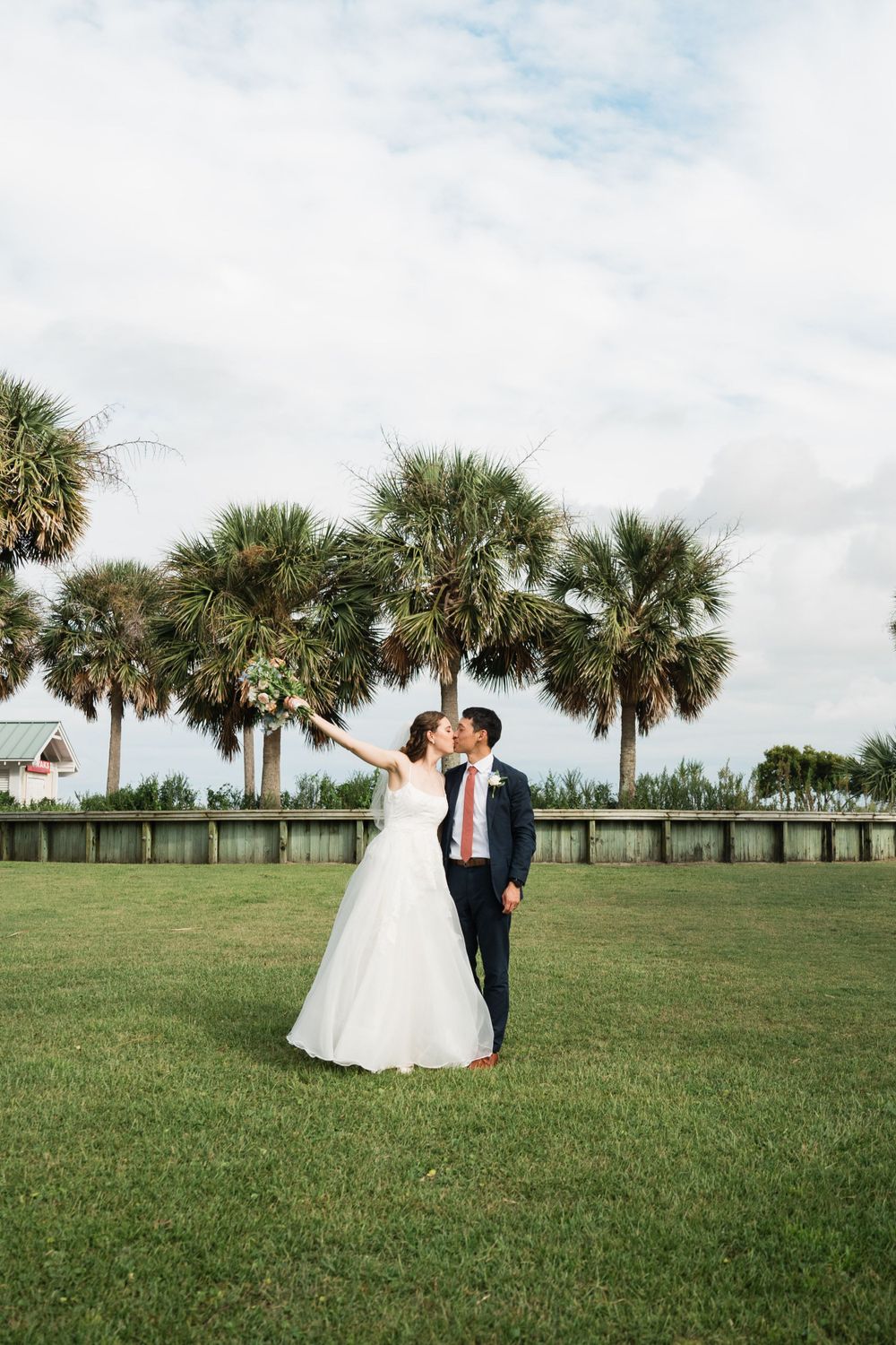 Wedding couple embraces on a green lawn with tall palm trees and cloudy sky in the background.