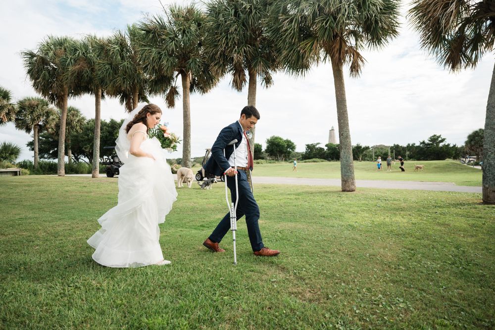 Bride in flowing white dress and groom walk across a grassy lawn lined with palm trees.