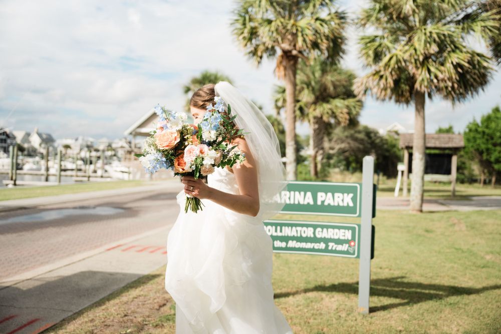 Bride with bouquet stands near a park sign surrounded by palm trees on a sunny day.