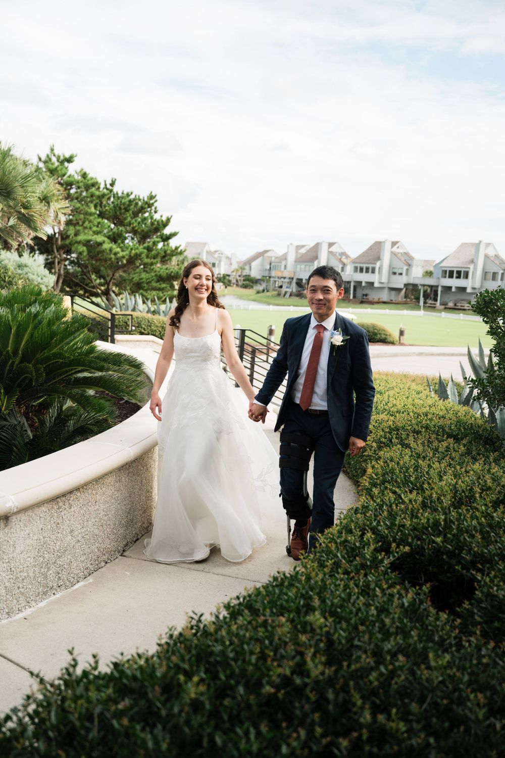 Wedding couple walking along coastal path with beach houses and tropical landscaping.