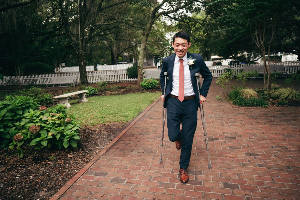 A groom walks alone on a brick path surrounded by greenery and white picket fences.