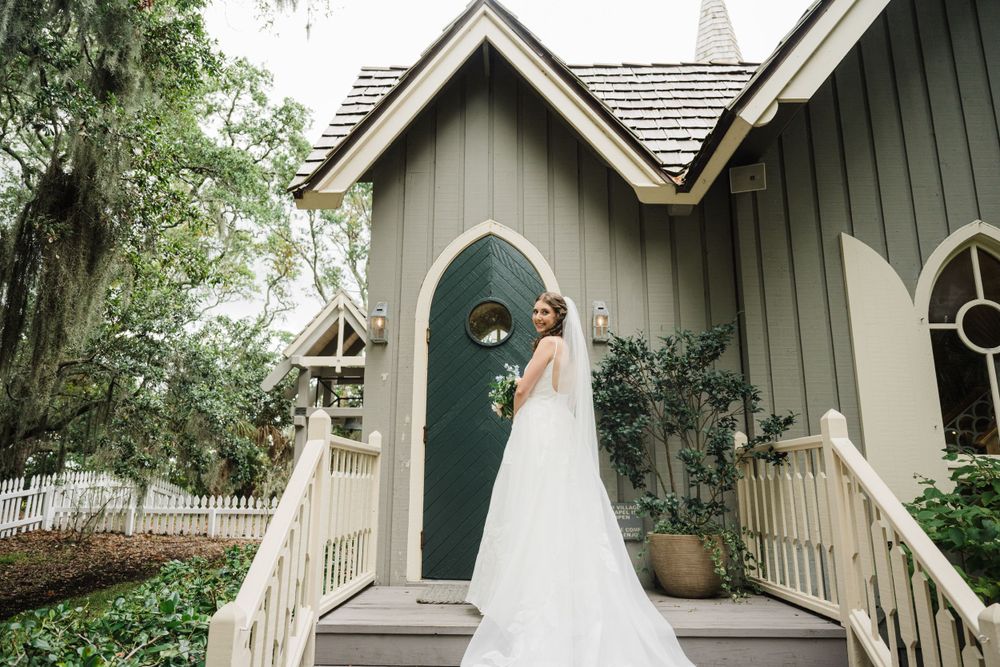 A traditional white chapel with grey siding and a teal door features a covered front porch and ornate gothic windows.
