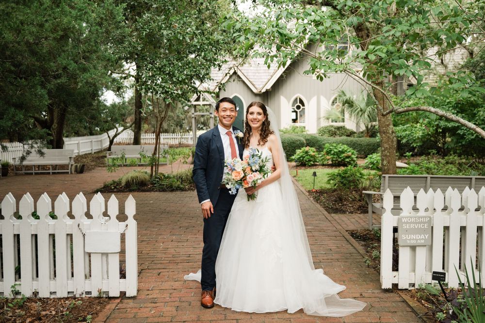 Beautiful wedding portrait in front of a quaint chapel with white picket fence and lush garden setting.