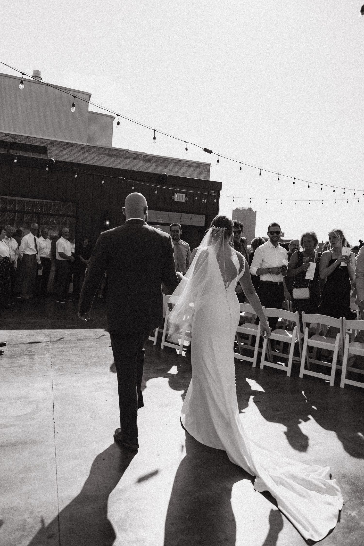 A black and white wedding ceremony photo taken on a rooftop with string lights and guests seated in white chairs.