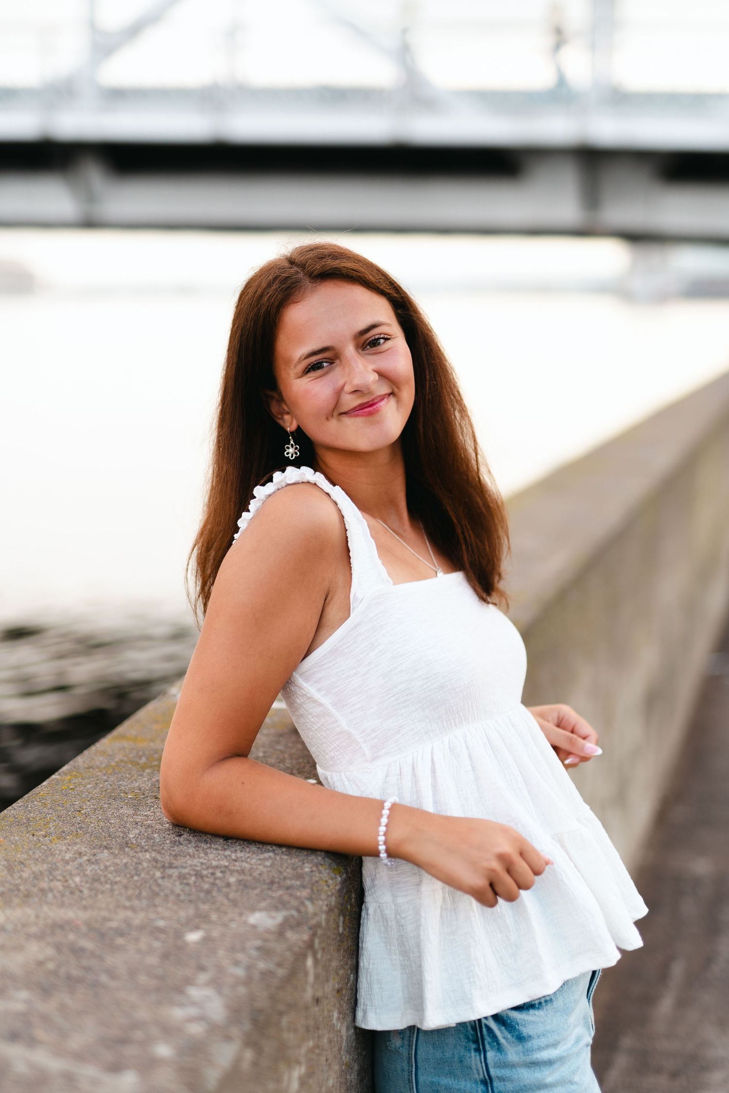 Young woman in white sleeveless top and jeans poses casually against concrete wall outdoors.