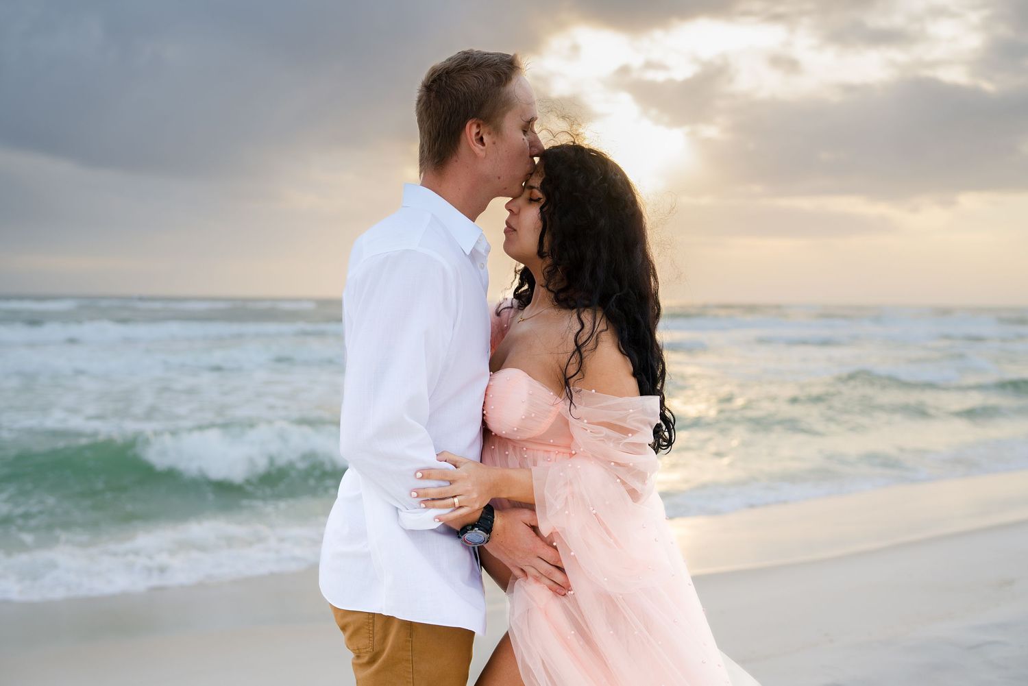 A romantic couple embraces on a beach at sunset with crashing waves and cloudy skies in the background.