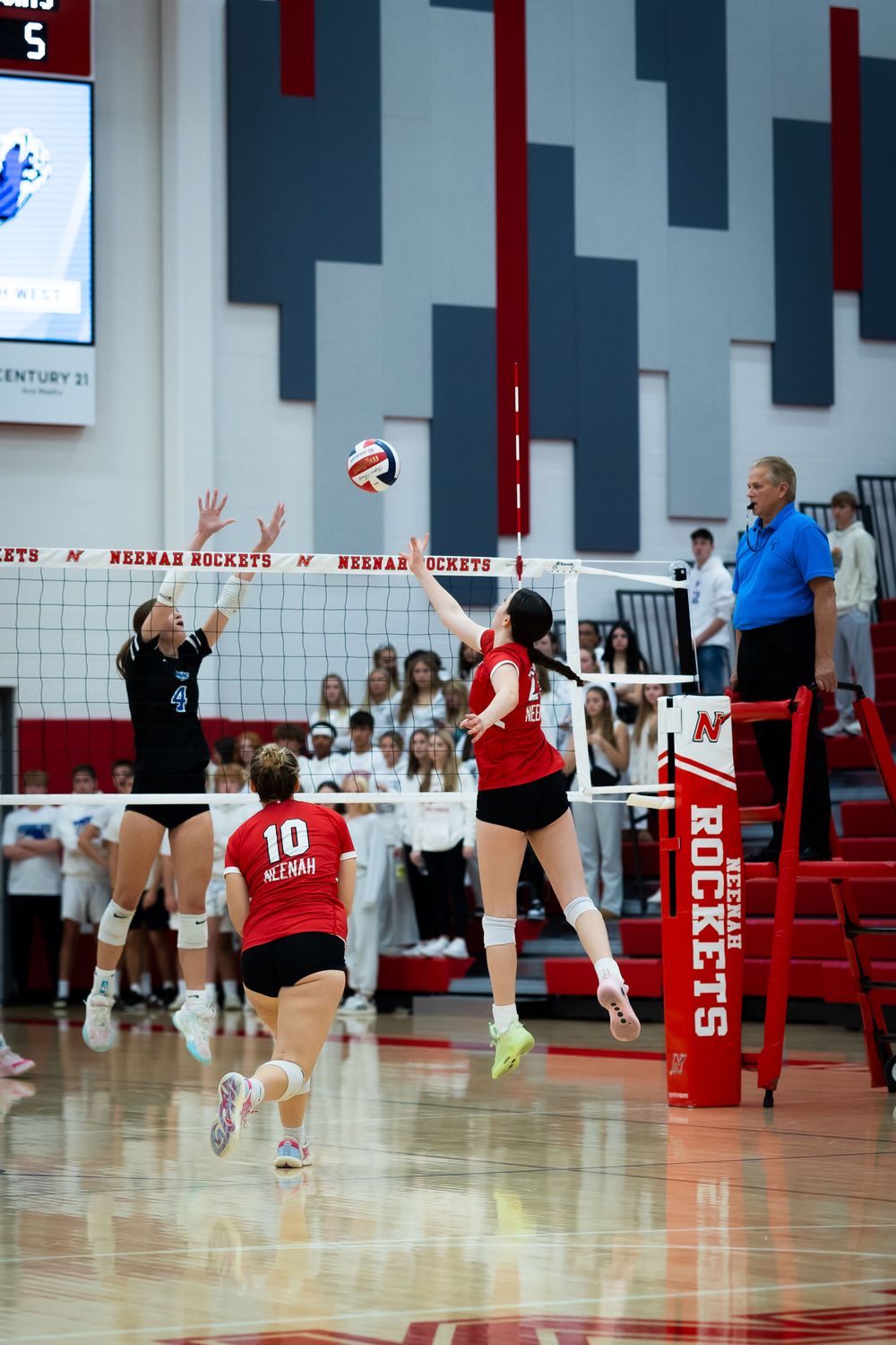 Volleyball players competing at net during indoor game with red and white court colors.