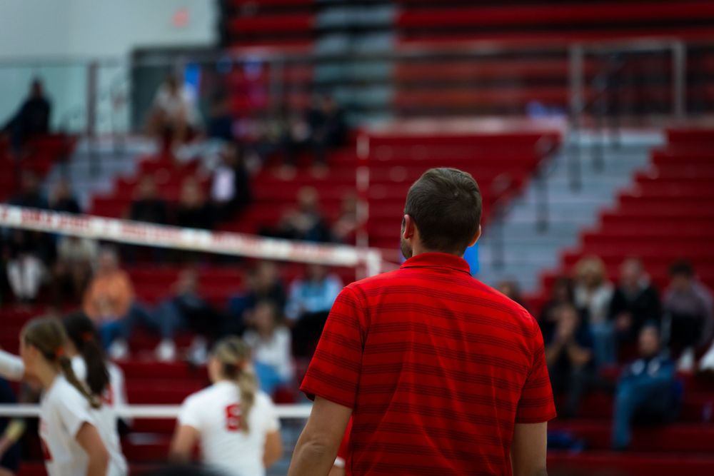 Coach in red shirt watches volleyball match from sideline.
