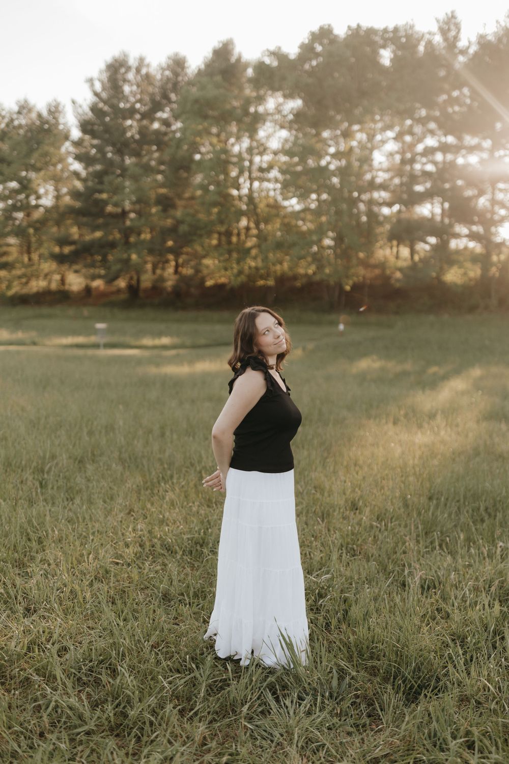 Figure in black top and white skirt standing in a meadow during golden hour. Virginia graduation/senior session.