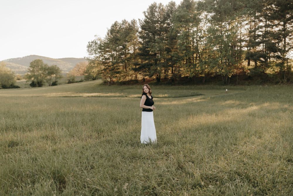 Distant figure in black and white outfit standing in an open field with pine trees in background. Virginia graduation/senior session.