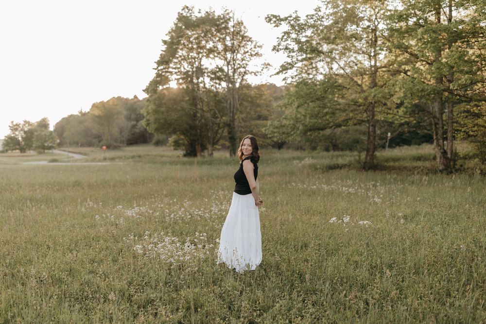 A full length portrait of a woman wearing a flowing white skirt standing in a golden summer meadow.  Virginia graduation/senior session.