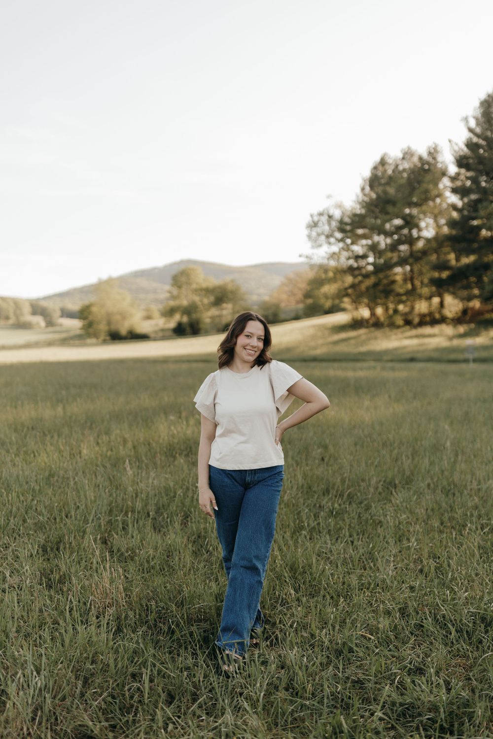 A portrait of a figure in a white t-shirt and jeans standing in a grassy field with mountains in the background. Virginia graduation/senior