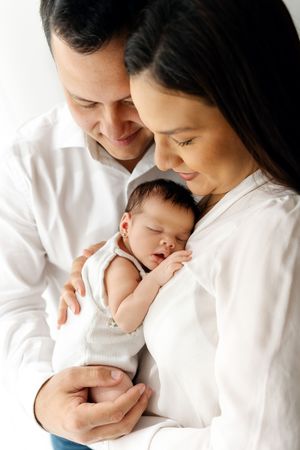 Tender moment as parents in white shirts embrace their sleeping newborn baby against white background.