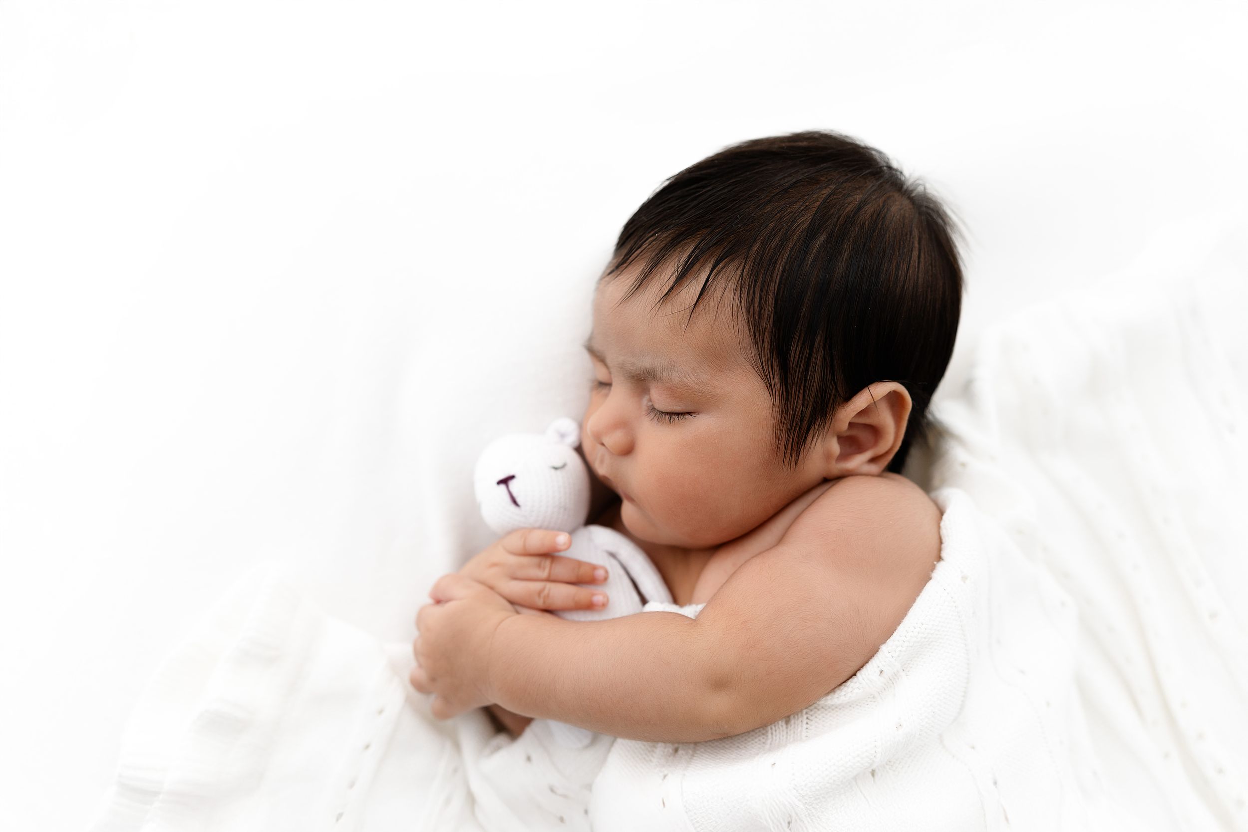 Newborn baby sleeping peacefully on white bedding while hugging a small stuffed animal.