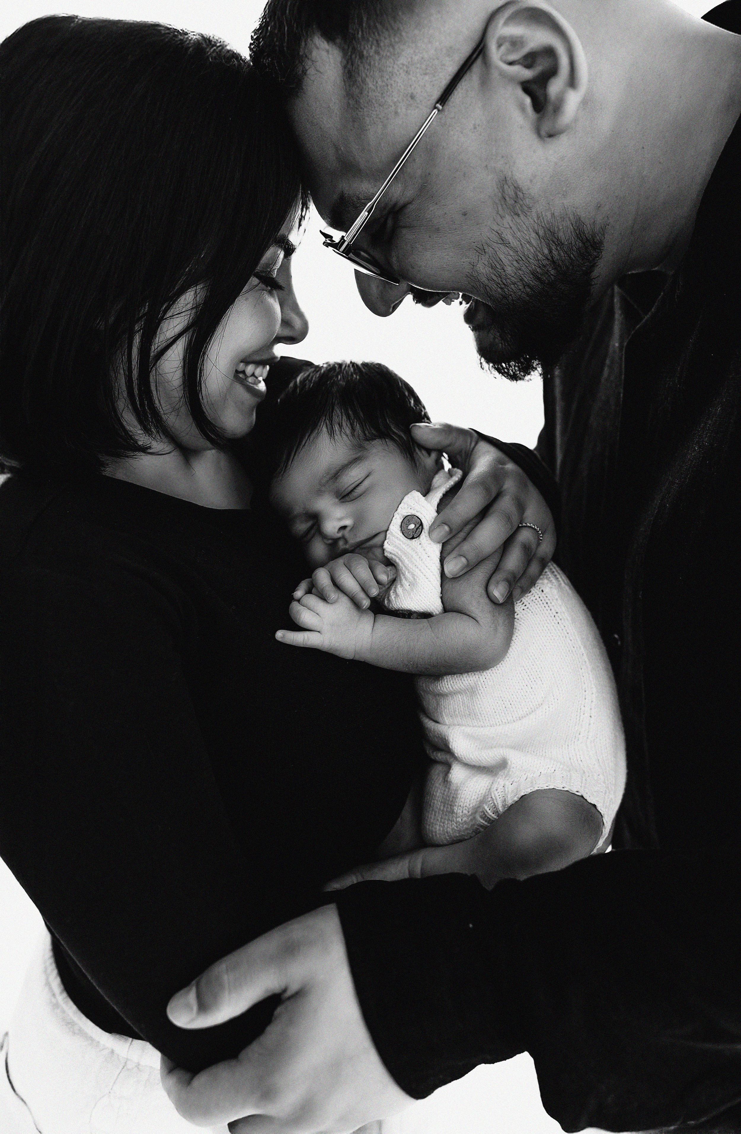 A black and white intimate family portrait shows parents cradling their sleeping baby in a tender moment.