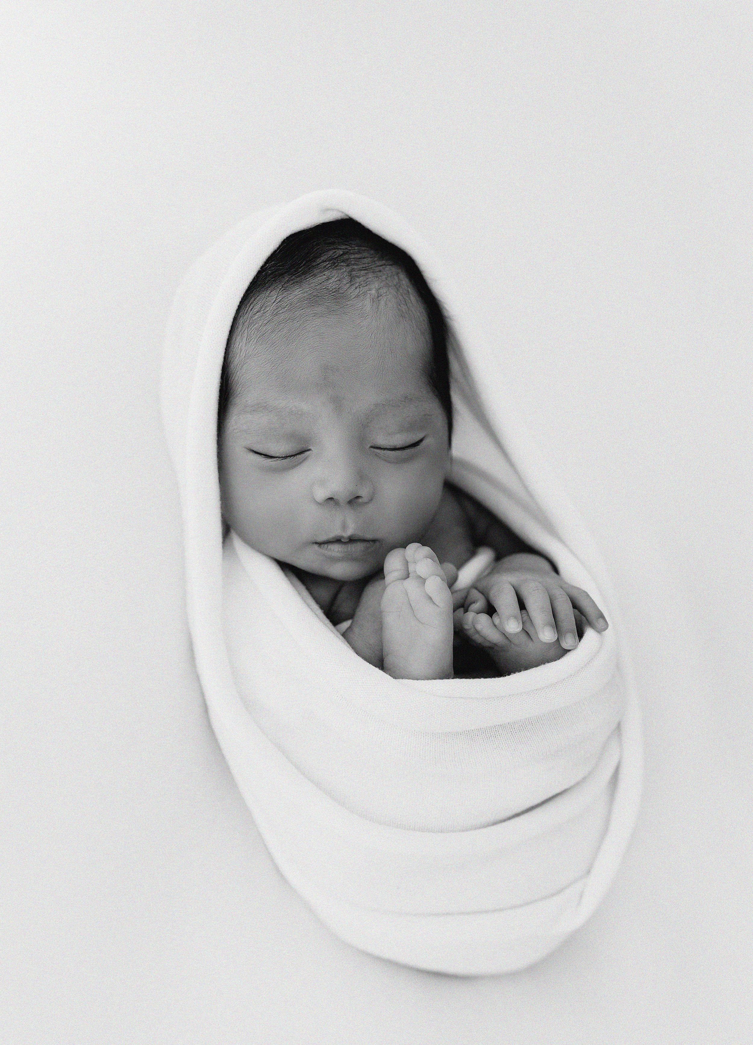 Black and white photo of a peaceful sleeping newborn baby swaddled in soft white blanket against light background.