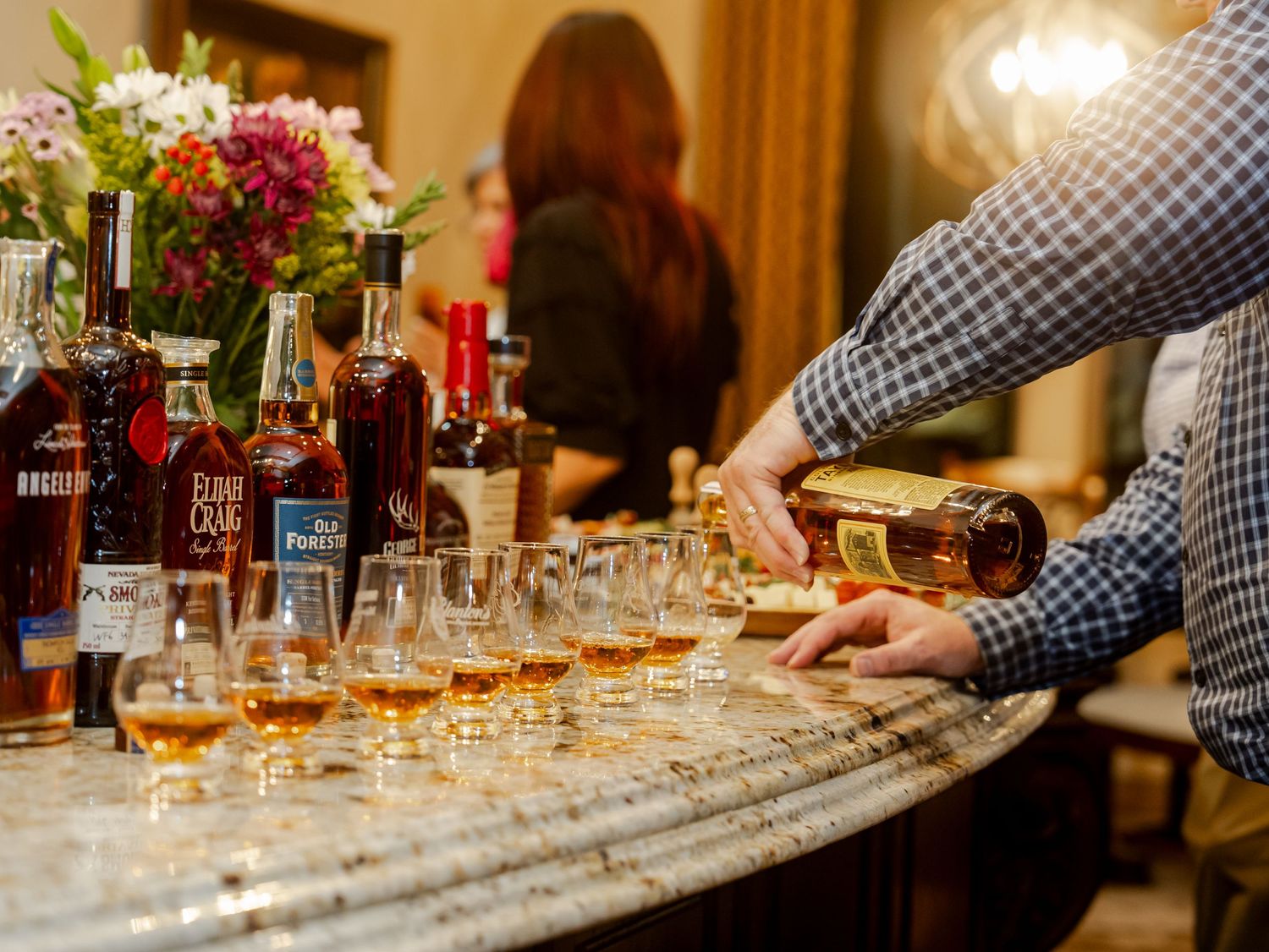 A row of bourbon bottles and whiskey glasses arranged on a marble bar counter during a tasting event.
