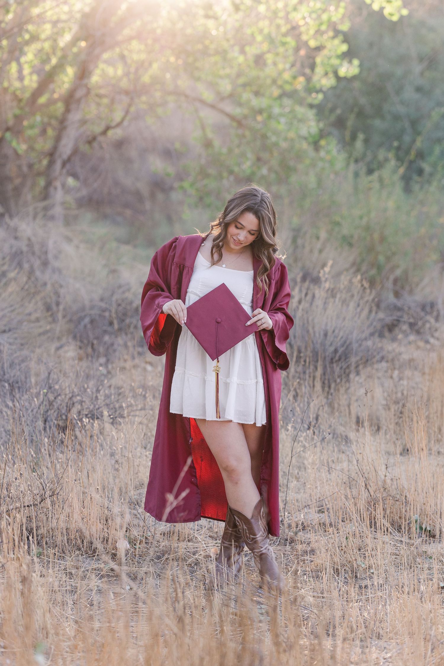 A graduate in a white dress and burgundy graduation gown looks down at their diploma while standing in a golden field.