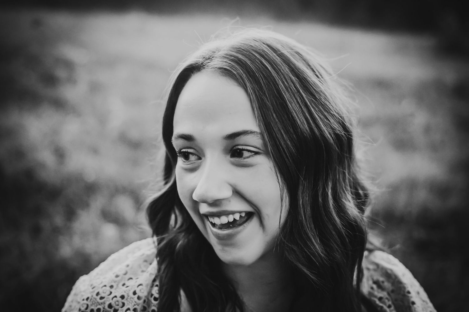 Black and white portrait of someone with wavy dark hair laughing while looking to the side in an outdoor setting.