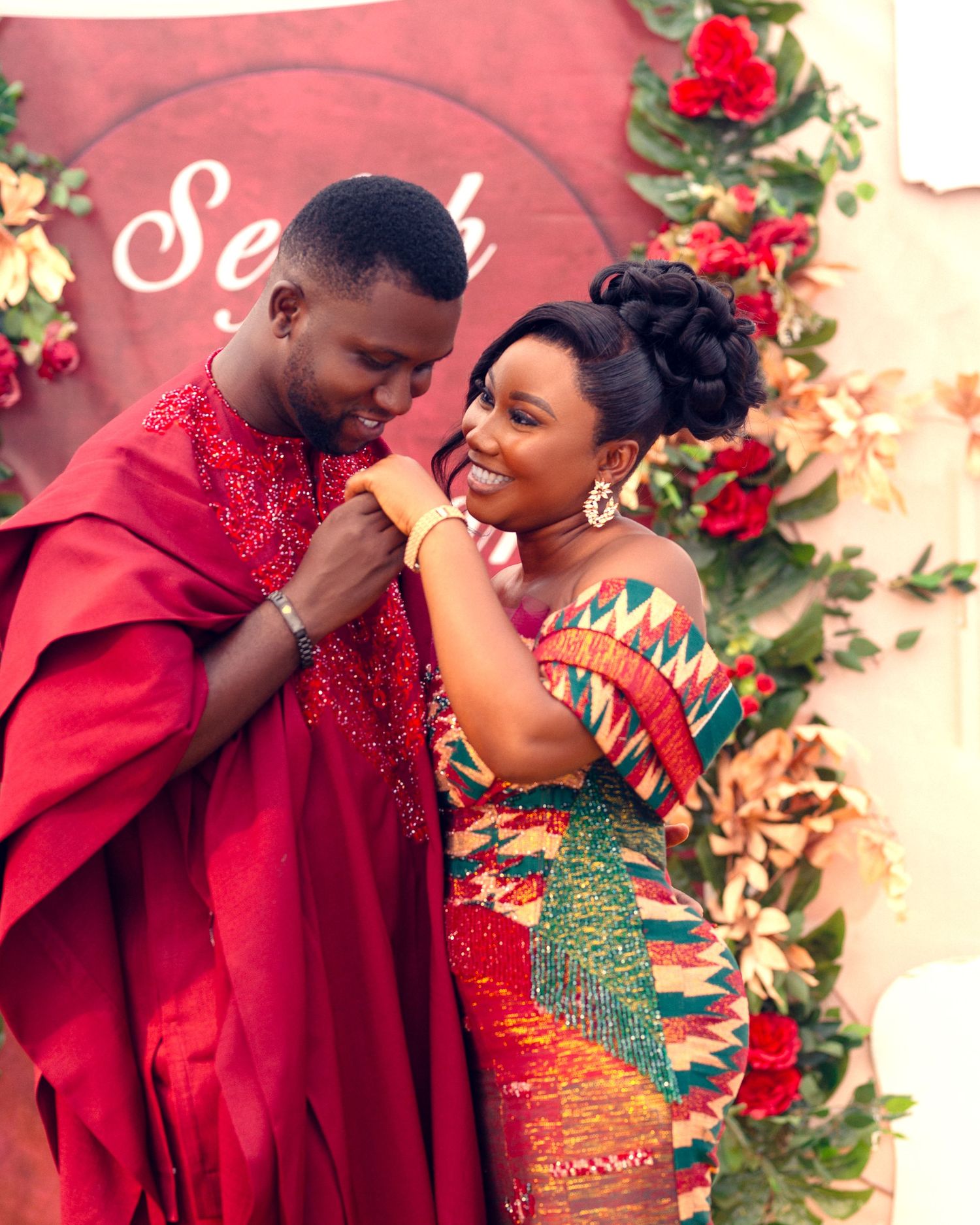 Romantic couple embraces against floral backdrop wearing traditional African fashion in red and print patterns.