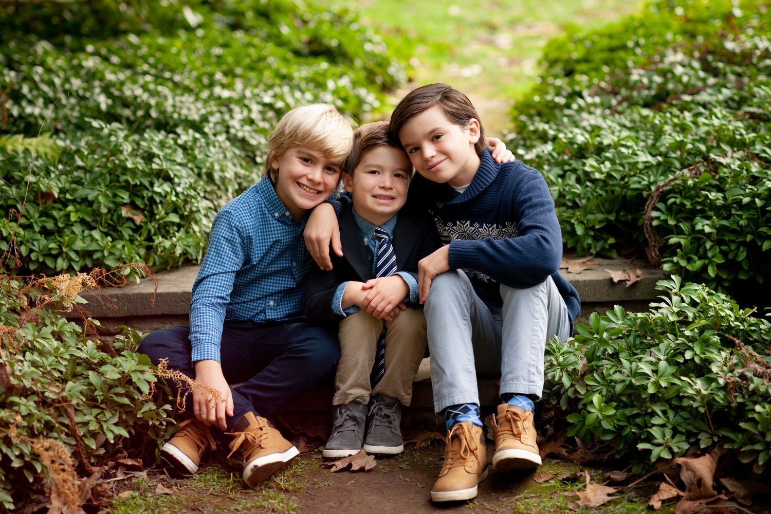 Three young boys smiling and posing together on garden steps surrounded by lush green foliage.