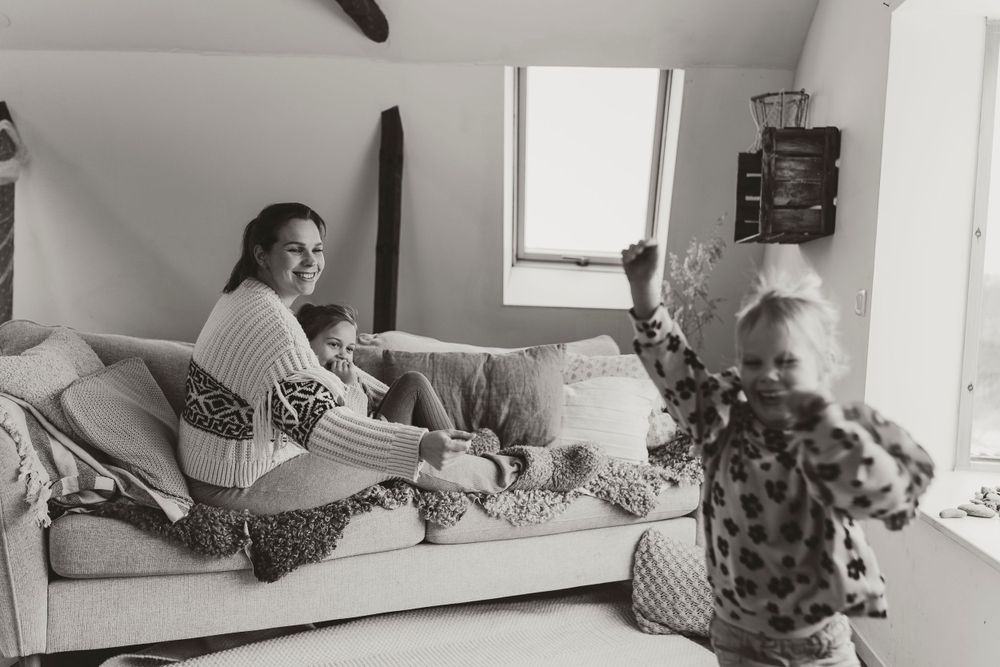 Black and white photograph showing a casual family moment at home on a couch while a small child plays in the foreground. Fotografering