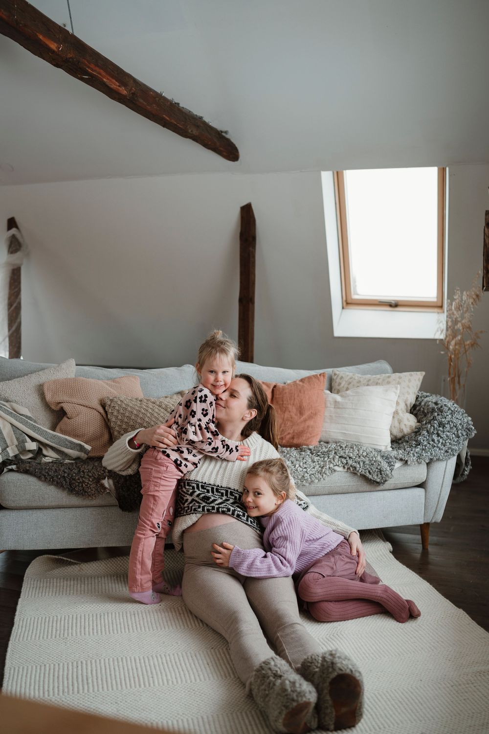 Cozy attic living room with exposed wooden beams, skylight, and family lounging on gray sofa with pink accents. Gravidfotografering Blekinge
