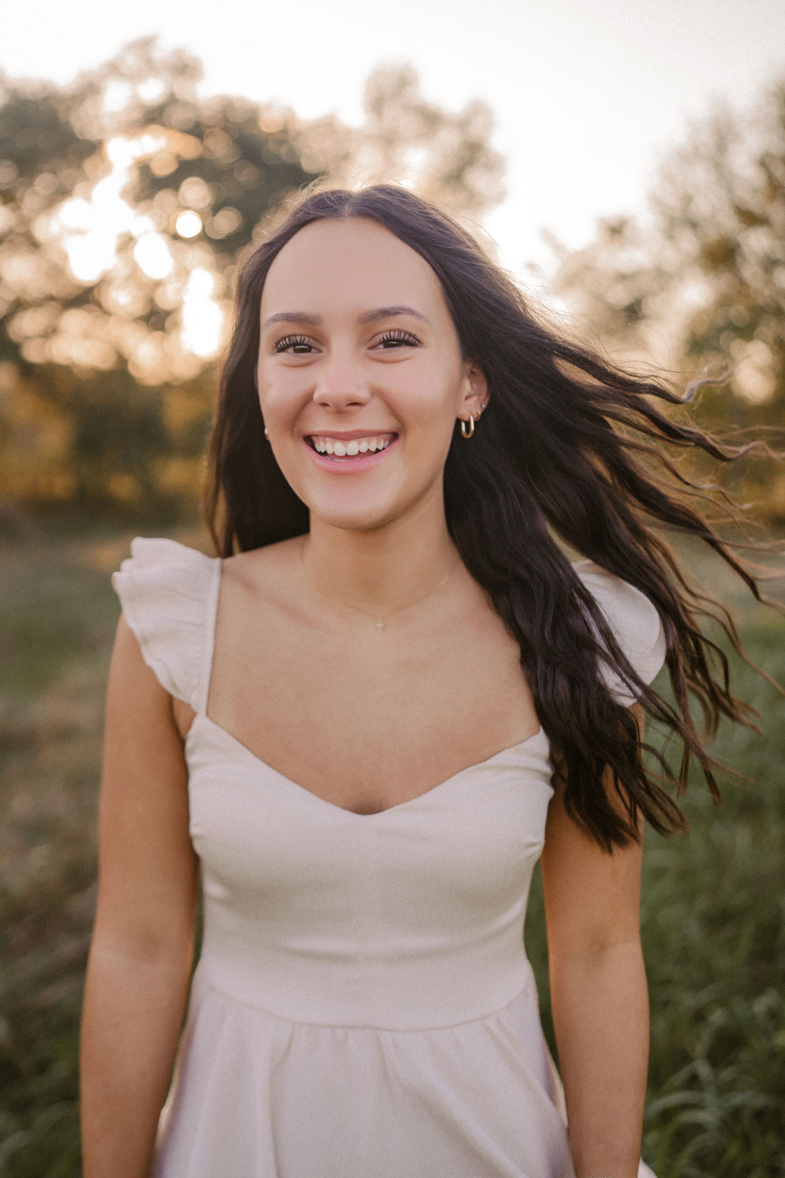 A smiling young woman with long dark hair wears a white summer dress while standing outdoors at golden hour.