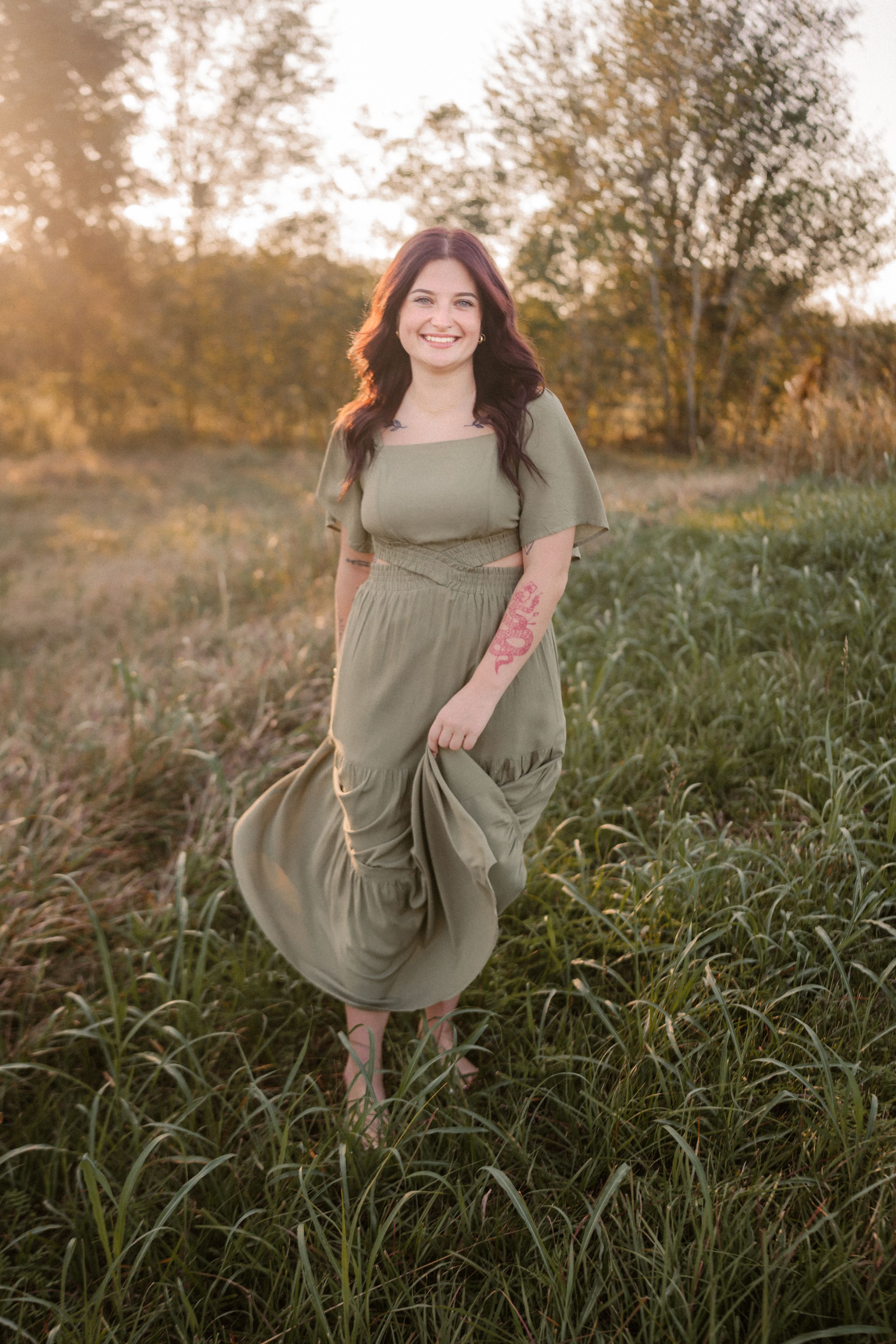 A person in an olive green dress stands in a sunlit field of tall grass at golden hour.